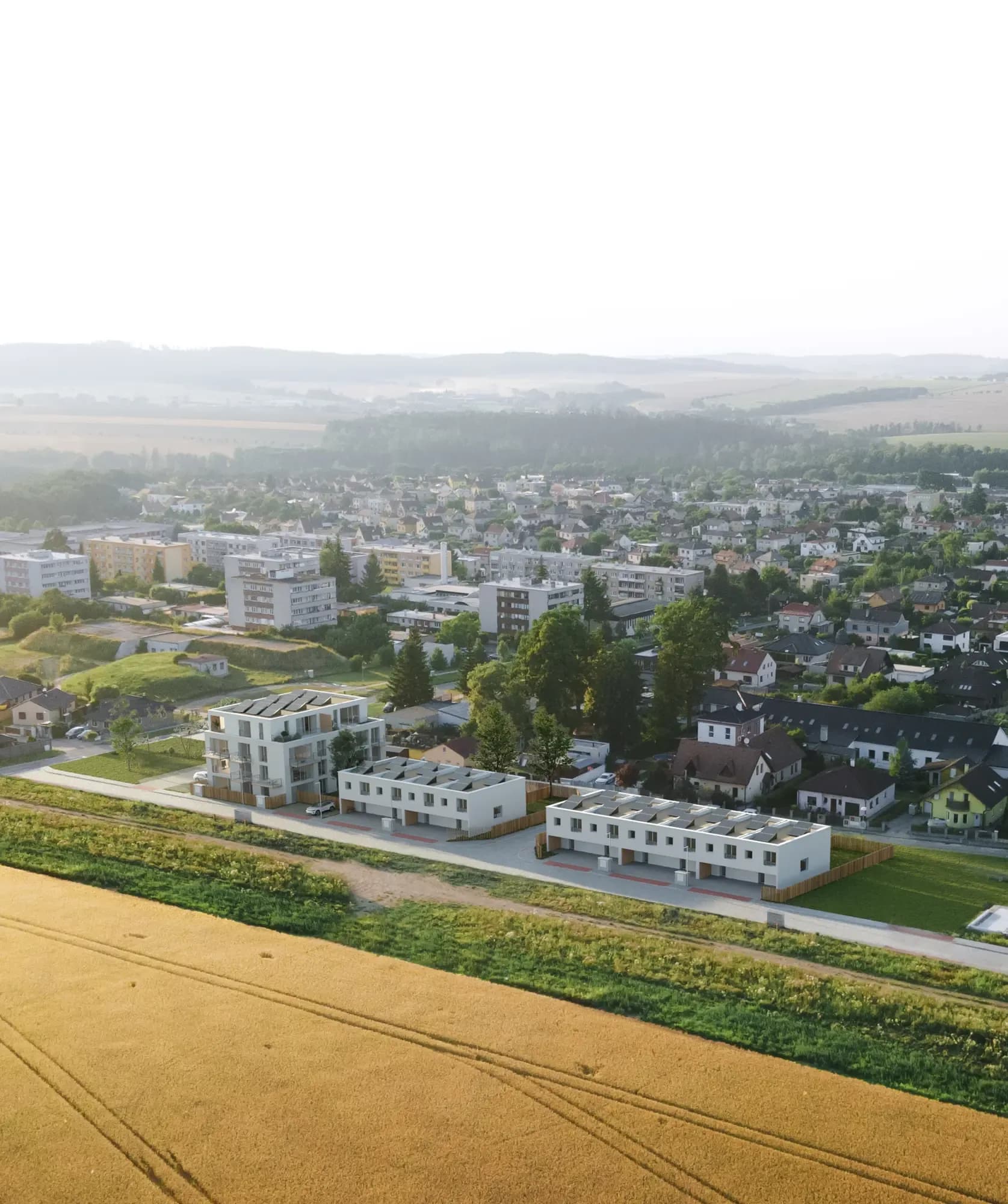 Letecký pohled na projekt Bydlení Vlašim při západu slunce – řadové domy, bytový dům a město v pozadí